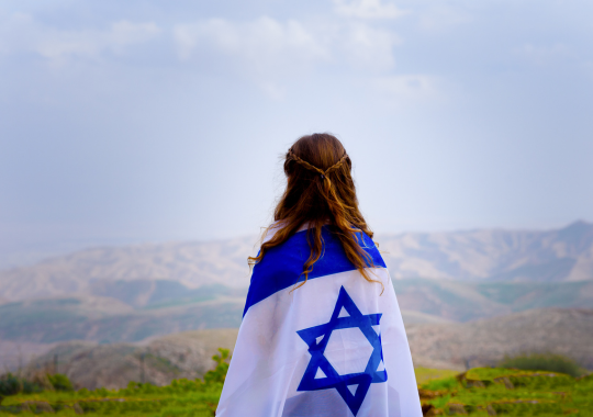 נערה עטופה בדגל ישראל ברקע הרים שמיים ומשטח ירוק של עשב